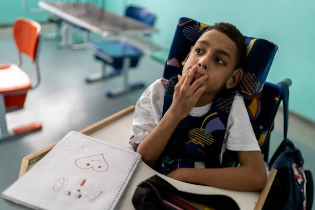 A boy in a wheelchair with head support sitting thoughtfully at his classroom desk